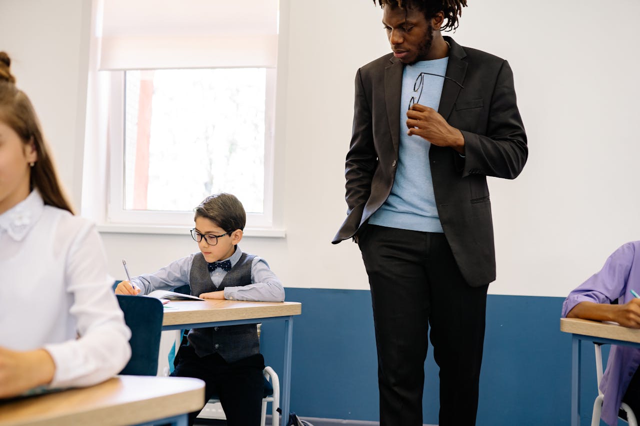 A teacher in a black suit supervises young students working at desks in a classroom.